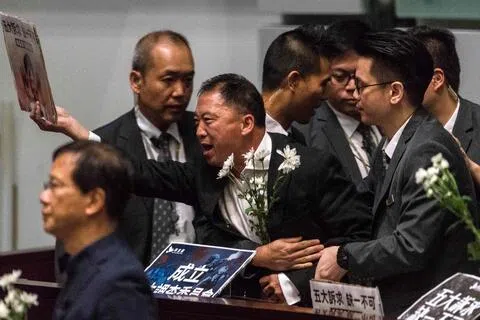 香港前民主党主席胡志伟原本被令遣返，后来获准在英国逗留半年。图为胡志伟（中）2019年在立法会质询时，向时任香港特首林郑月娥（不在图内）高举抗议标语。（法新社档案照）