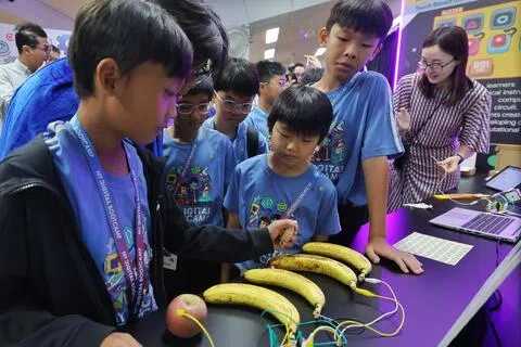 后港小学和宏茂桥小学的学生星期一在AI版数码学习营的启动日活动上，使用由键盘模拟器Makey Makey制作的水果“琴”。敲打香蕉或苹果会触发电脑编程，发出钢琴、吉他或其他乐器的声音。（唐家鸿摄）