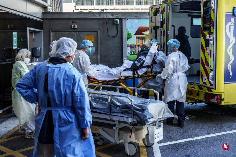 香港医护人员穿上全副个人防护装备，准备将一名年迈病人从救护车转移到伊丽莎白女王医院的急症室。旁边担架躺着一具不幸逝世的病人遗体，正等候被送入医院殓房。（彭博社）