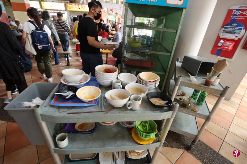 雅柏熟食中心的归还托盘处堆满碗碟，因为空间有限，不少食客只得将餐盘和餐具放置在一旁的推车上，导致推车上也堆积了不少脏碗碟。（梁麒麟摄）