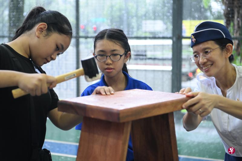 女少年旅成员和受益者一起组装凳子，再用锤子固定。这些家具之后会送到30户弱势家庭。（海峡时报）