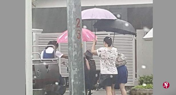 两名圣尼各拉女校的女生背着书包，由母亲撑伞，冒 雨帮老妇解困。（Stomp)