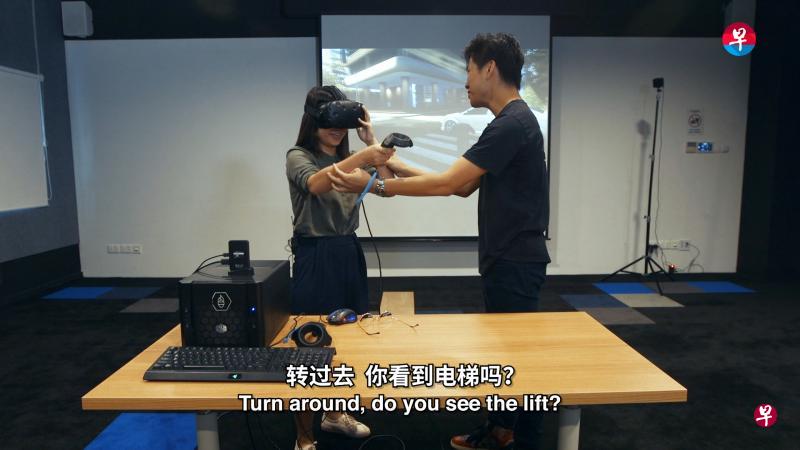 华文媒体集团新闻中心记者卓彦薇（左）在虚拟实境体验设计师丘勇凯（右）的指导下，体验虚拟实境游戏“Richies Plank”，“走进”电梯，感受高空俯瞰的特殊体验。（视频截图）