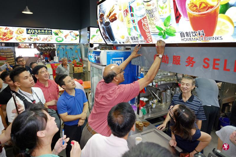 副总理兼经济及社会政策统筹部长尚达曼昨天为裕廊中央广场主持开幕仪式后，走访一楼的湿巴刹和咖啡店，并在一家茶水摊的招牌上签名留念。同行的还有社会及家庭发展部长李智陞（左三）和裕廊集选区议员洪维能（左二）。（曾坤顺摄）