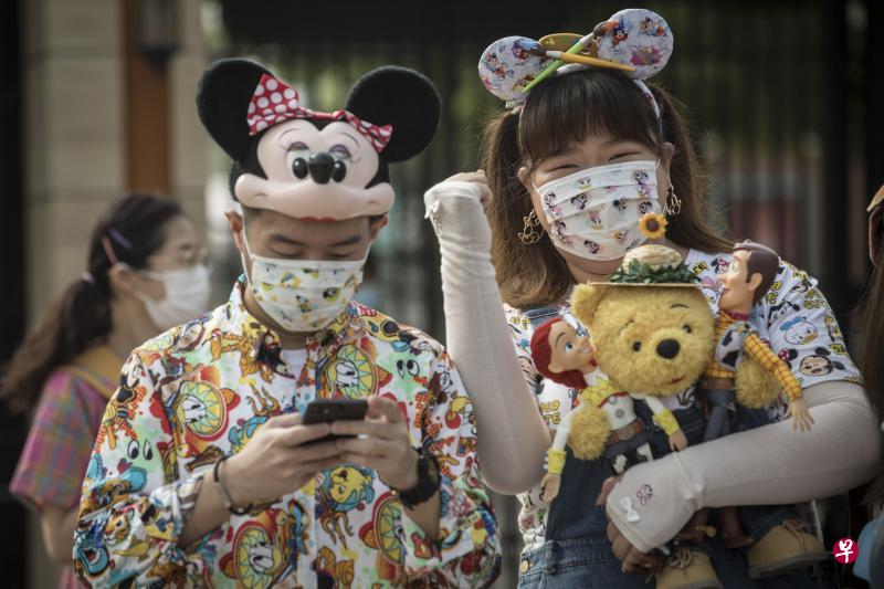 图中这对游客，从帽子发箍、口罩、手套到衣服，都离不开米老鼠主题。女生还兴奋地抱着小熊维尼和玩具总动员布偶，一起游园。（彭博社）