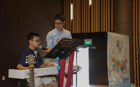 担任导师的张乐声（右）在学生钢琴演奏时协助举麦克风，学生在台上完成自弹自唱的表演。（陈心媛摄）