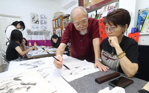 新加坡美术总会前会长梁振康（持画笔者），在丙午马年到来前，指导学员潘少琼画马。（郑一鸣摄）